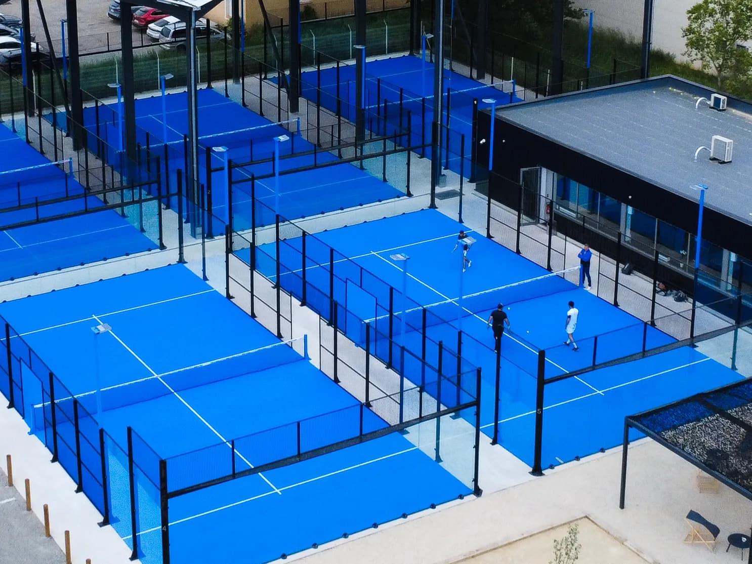 Four blue padel courts from the air, players on court and the clubhouse alongside